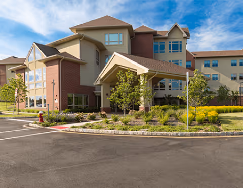 Exterior view of a multi-story senior living facility building with a covered entrance, surrounded by landscaped greenery and a clear blue sky.