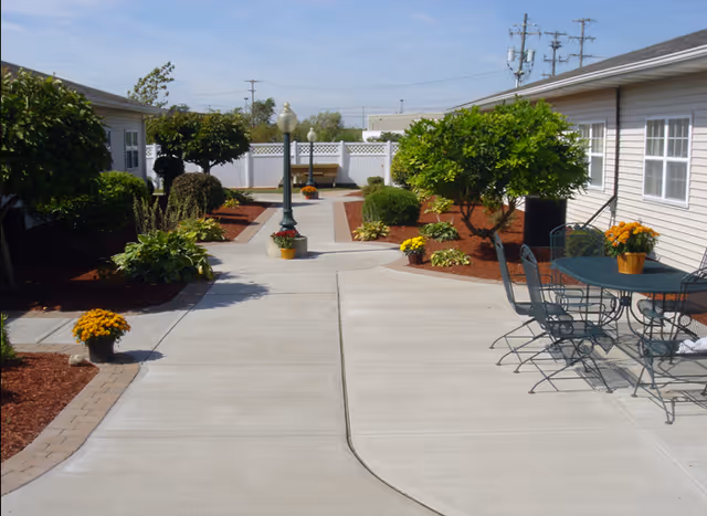 Sunny landscaped courtyard between single-story buildings with a concrete walkway, trees and shrubs, a lamppost, and a patio table with chairs.