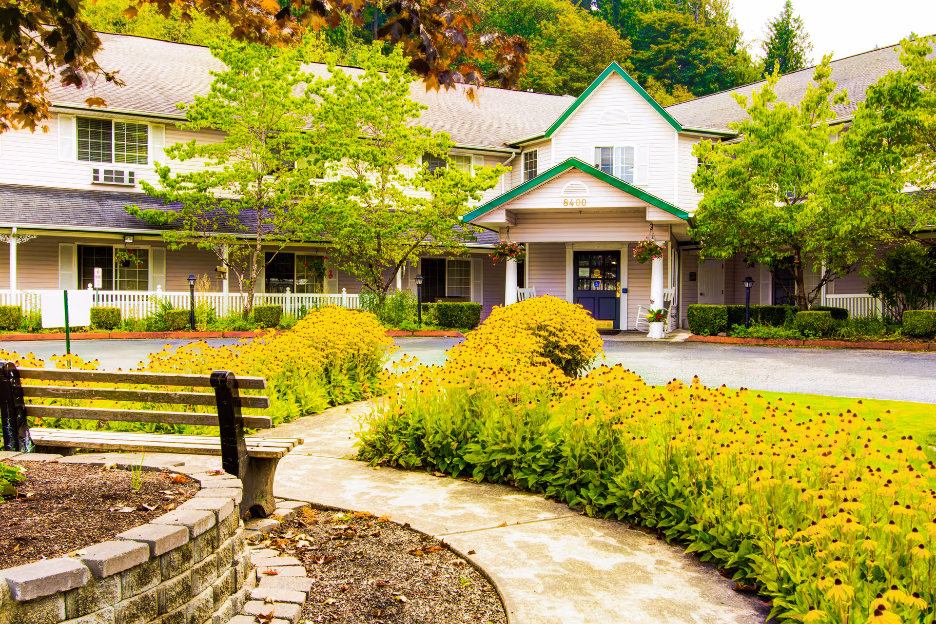 Front entrance of a senior living building with a curved walkway, bench, and beds of yellow flowers.