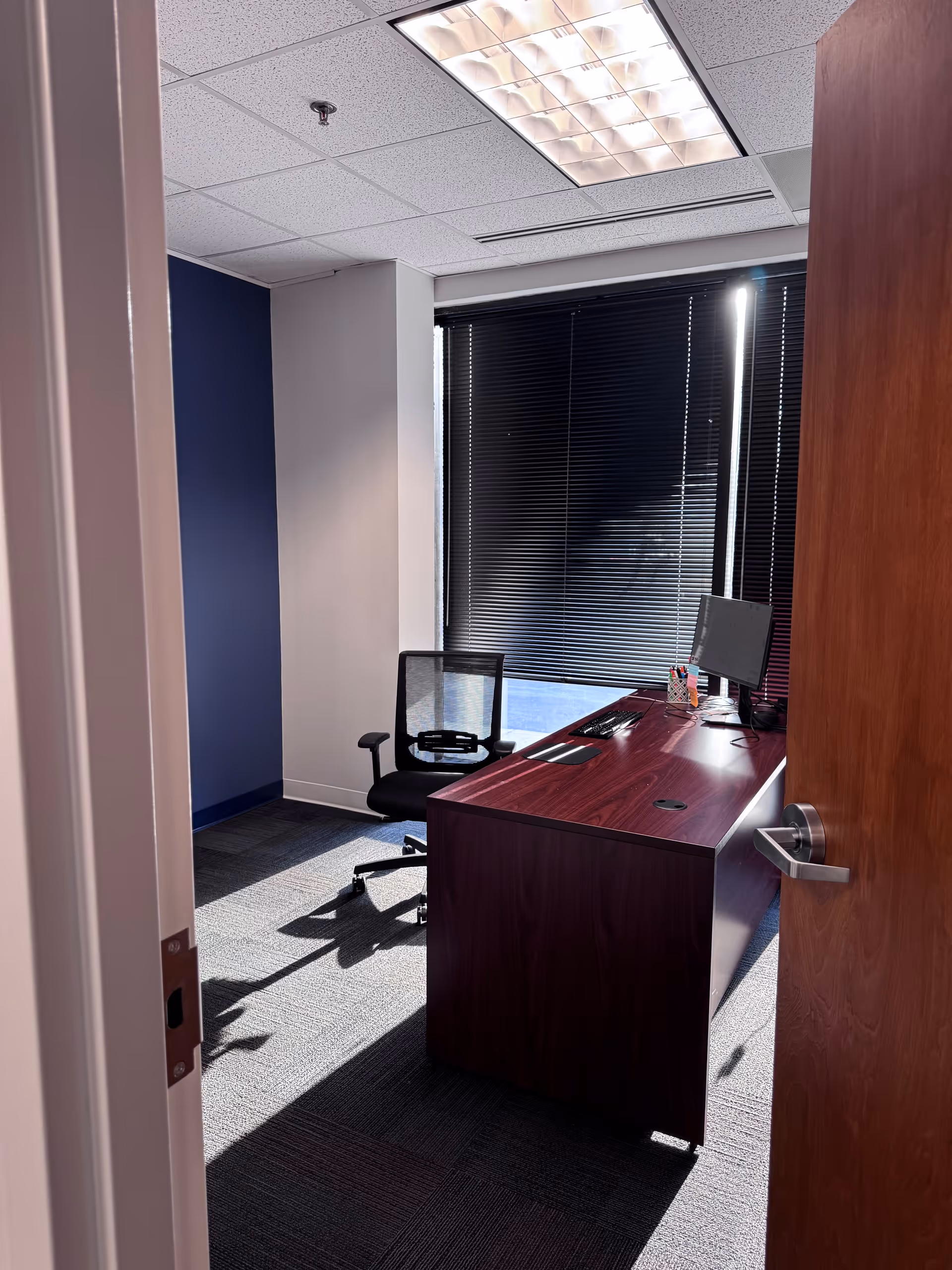Small office with a wooden desk, chair, computer monitor, and dark blinds.