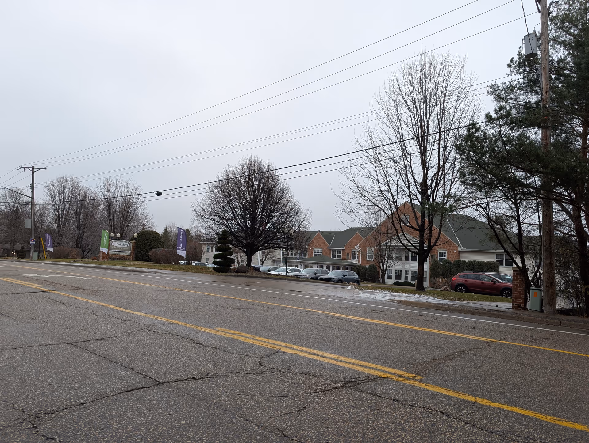 Exterior view of Heathers Manor senior living facility on a cloudy day, showing a brick building with multiple windows, a parking lot with cars, leafless trees, and a sign with the facility name near the road.