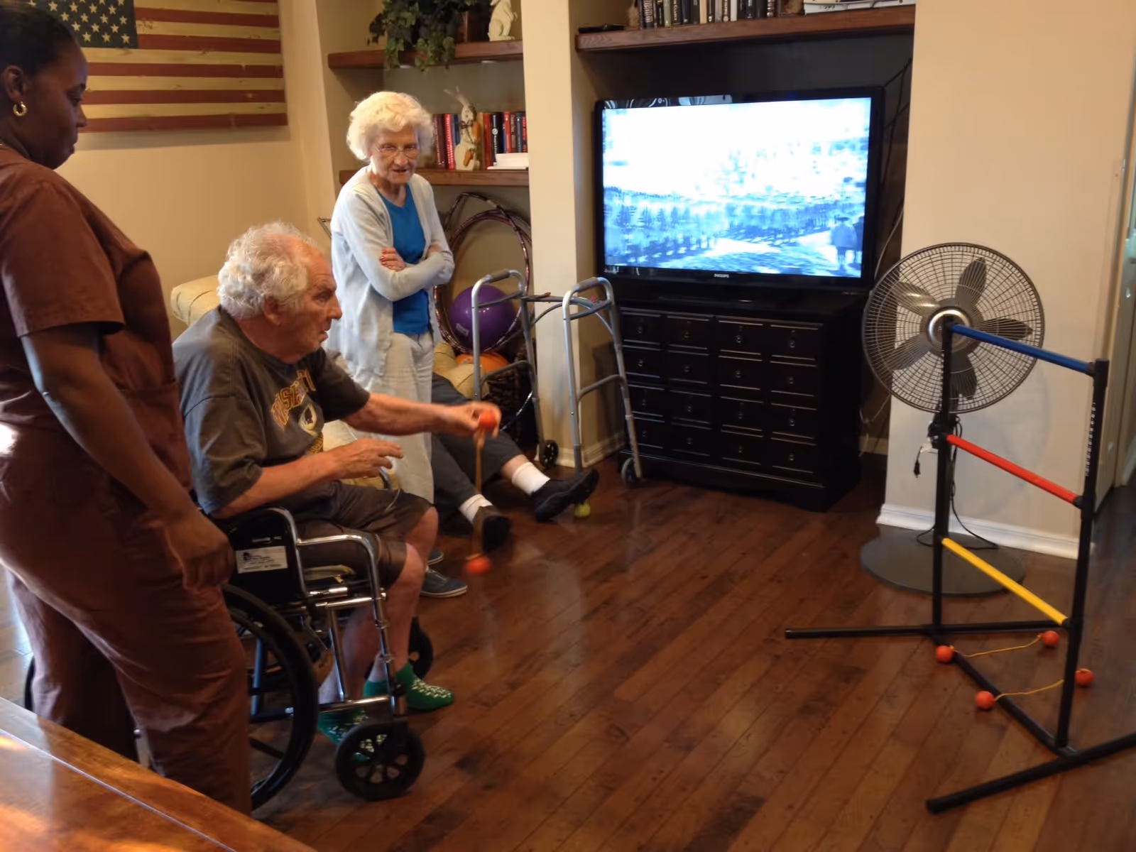 An elderly man in a wheelchair playing a ball toss game indoors, assisted by a caregiver standing beside him. An elderly woman stands nearby watching, with a walker and exercise balls in the background. A television and a standing fan are also visible in the room.