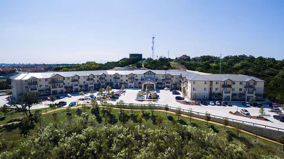 Aerial view of a three-story beige senior living building with balconies, a central entrance, a parking lot, and surrounding trees and landscaping.