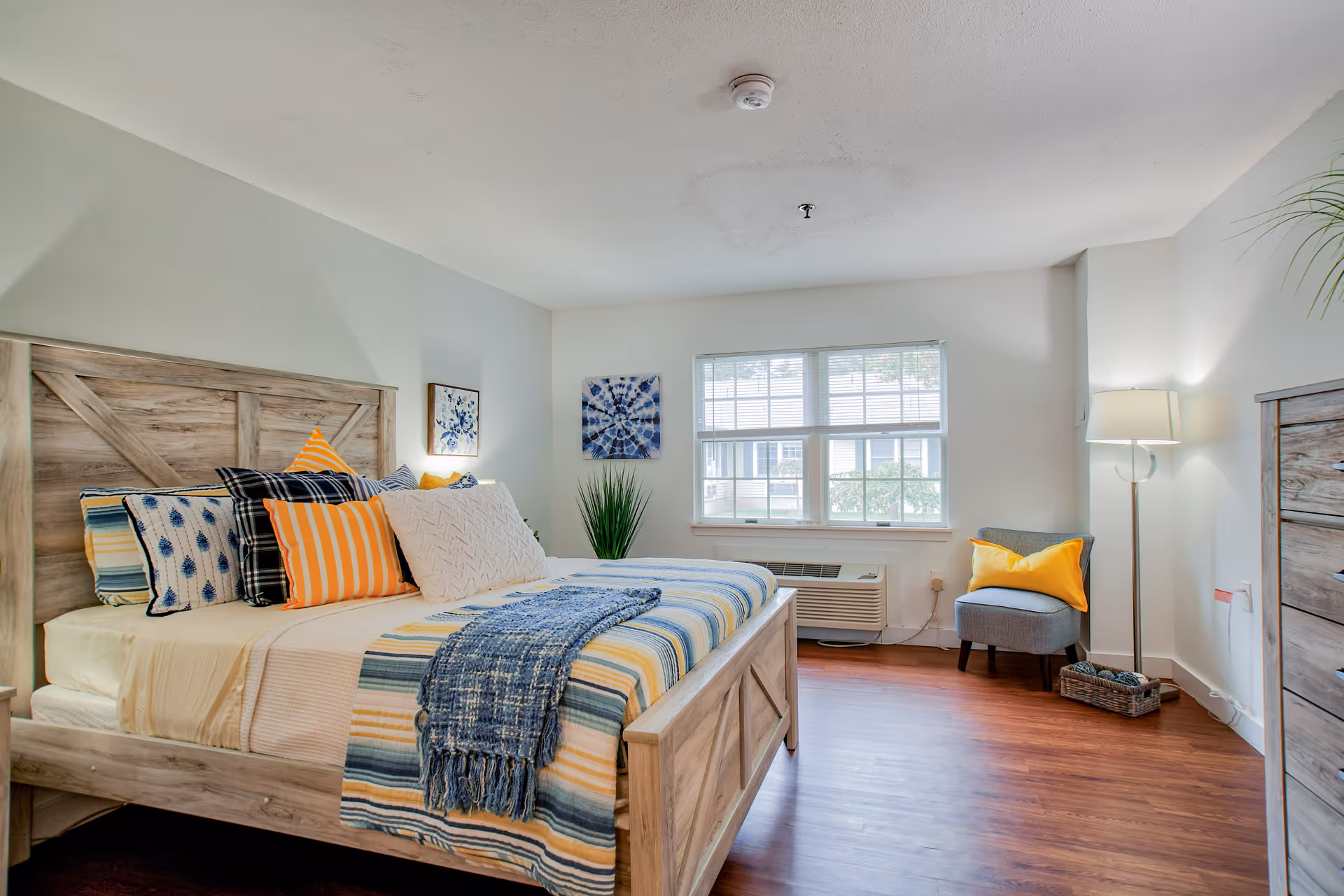 A cozy bedroom with a wooden bed, colorful pillows, window, and seating area.