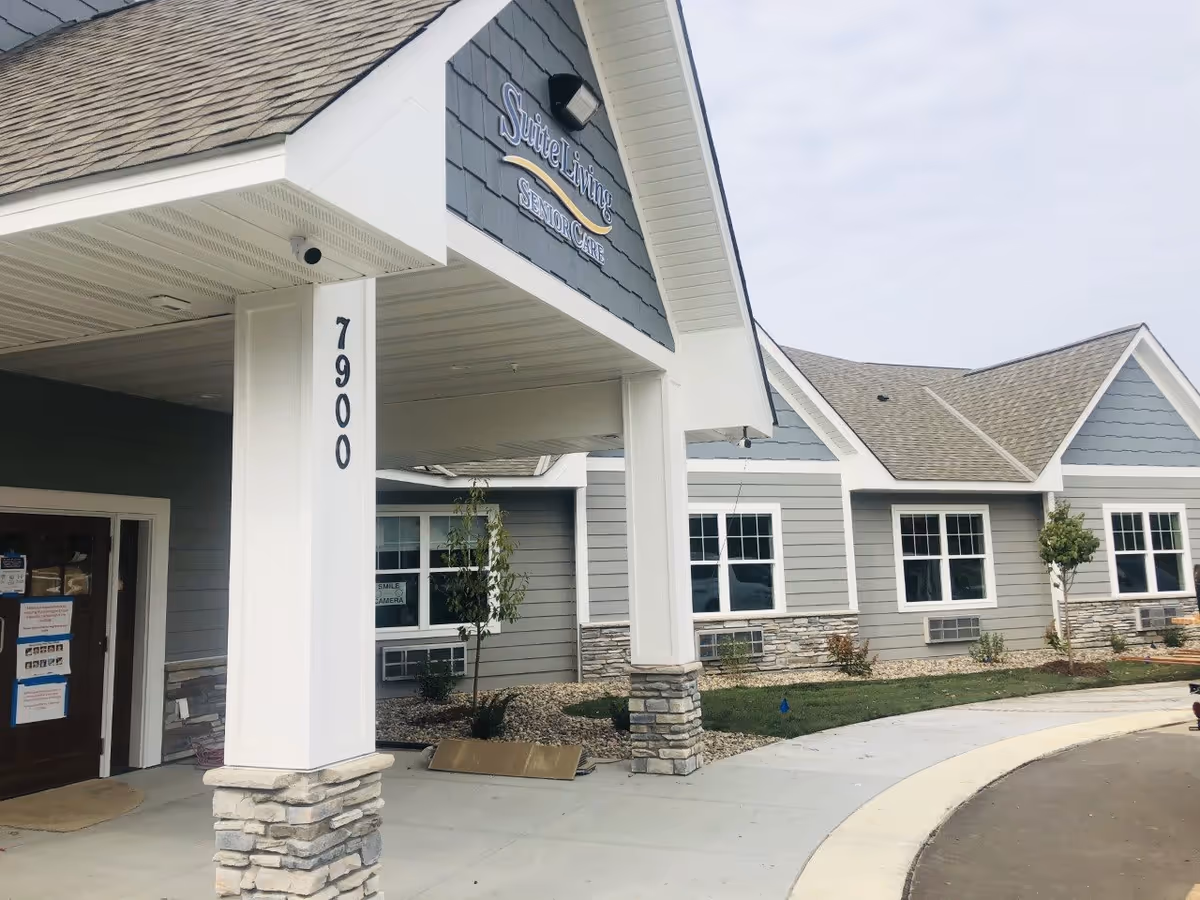 Exterior view of Suite Living of Inver Grove Heights senior care facility showing the entrance with a covered porch supported by white columns with stone bases, grey siding, multiple windows, and a curved driveway.