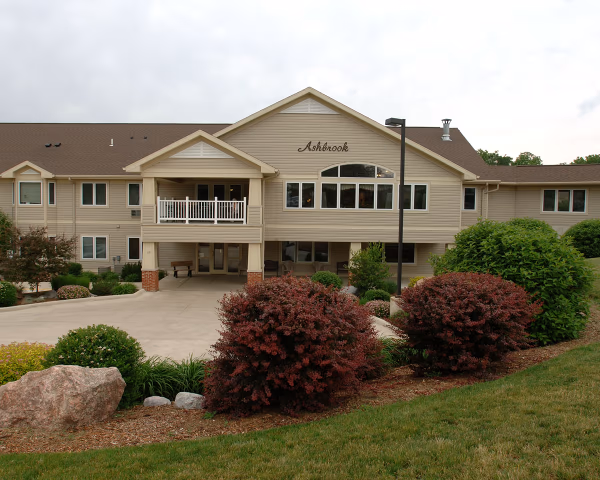 Front exterior of a beige two-story senior living building with landscaped shrubs and a driveway, sign reading "Ashbrook" above the entrance.