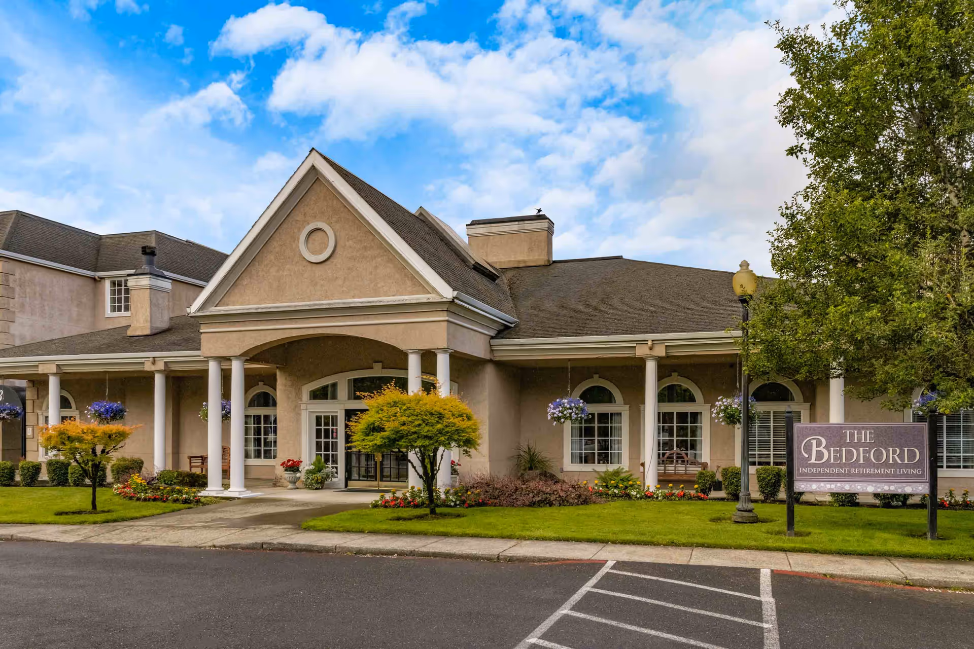 Front entrance of a senior living facility with a columned portico, manicured lawn, flower beds and a sign on the lawn.