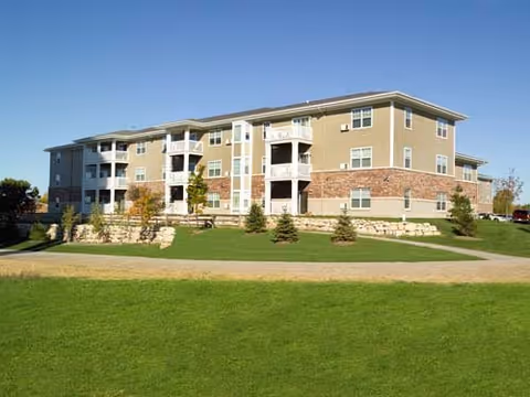 Three-story beige apartment-style senior living building with balconies, stone accents, and a manicured lawn under a clear blue sky.