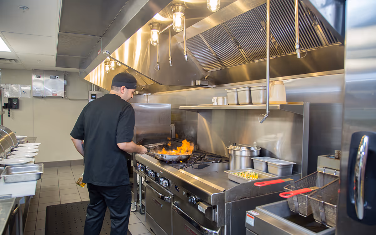 Chef in a stainless steel commercial kitchen, cooking at a large stove under overhead vents.