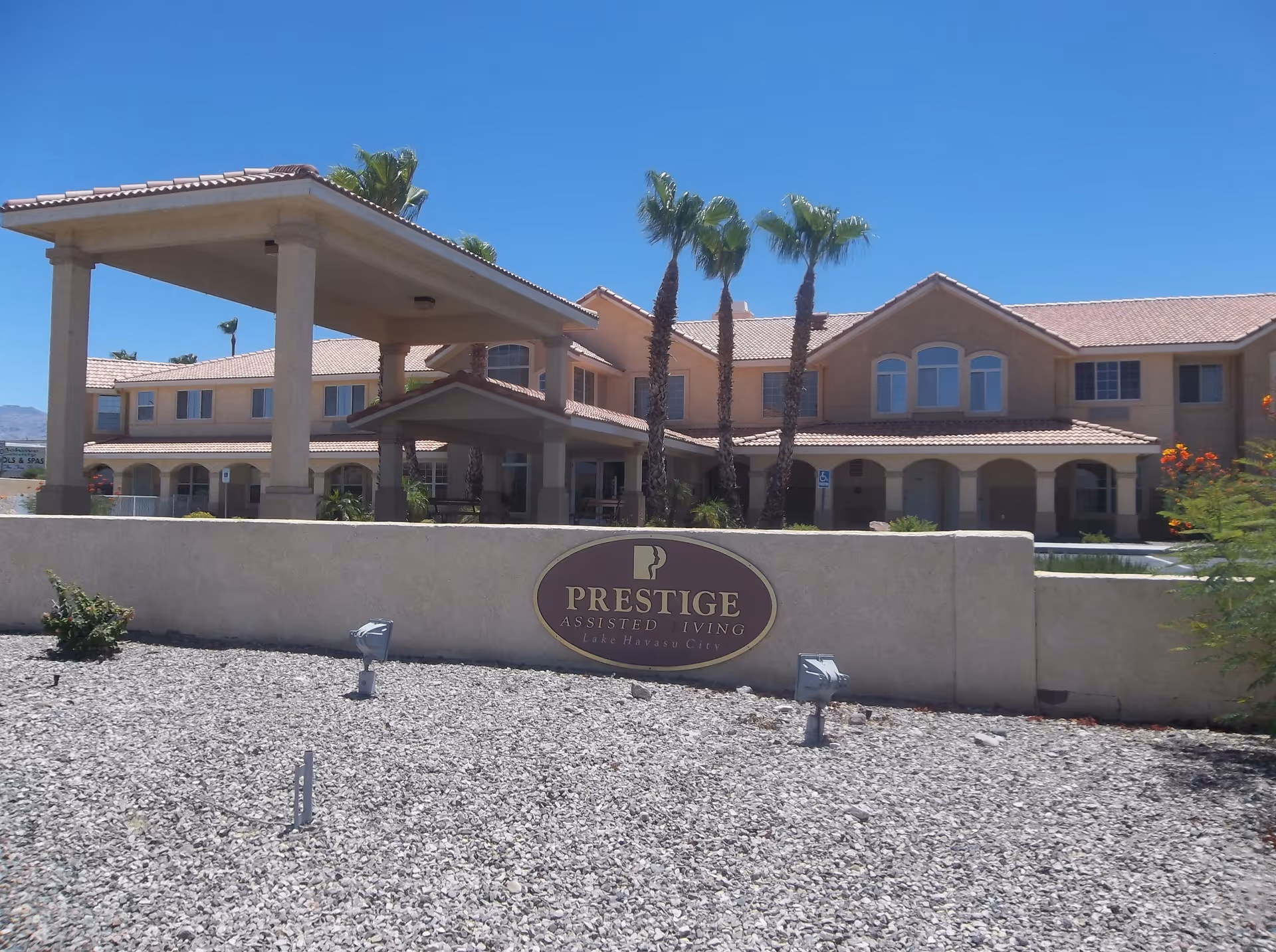 Exterior view of Prestige Assisted Living facility in Lake Havasu City, showing a two-story building with a covered entrance, palm trees, and a stone-covered front yard under a clear blue sky.