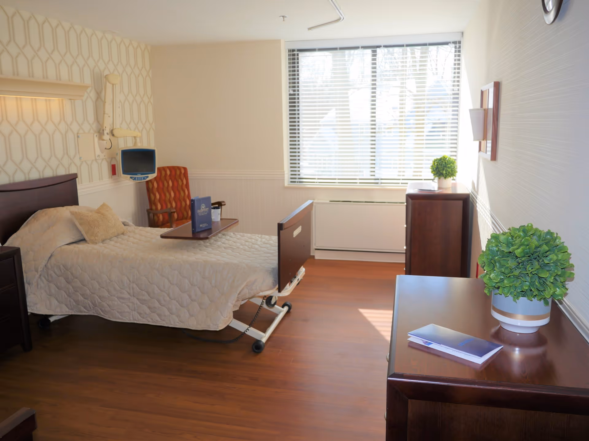 Single hospital-style patient bedroom with a bed, dresser, armchair, and large window.