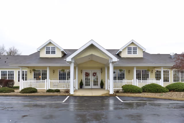 Front exterior view of a single-story building with a covered entrance supported by white columns. The building is painted light yellow with white trim and has two dormer windows on the roof. There are bushes and small trees planted along the front, and a wet parking lot is visible in the foreground.