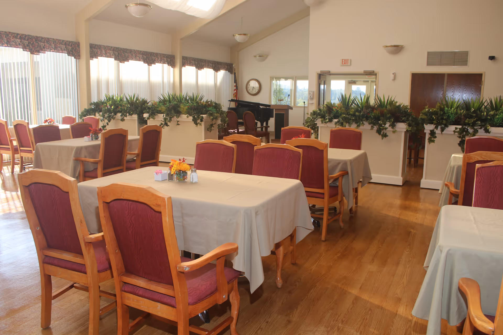A bright dining room with multiple tables covered in beige tablecloths and surrounded by wooden chairs with red cushions. The room has large windows with vertical blinds and floral valances, and there are green plants placed on white dividers. A piano and an American flag are visible in the background near the entrance door.