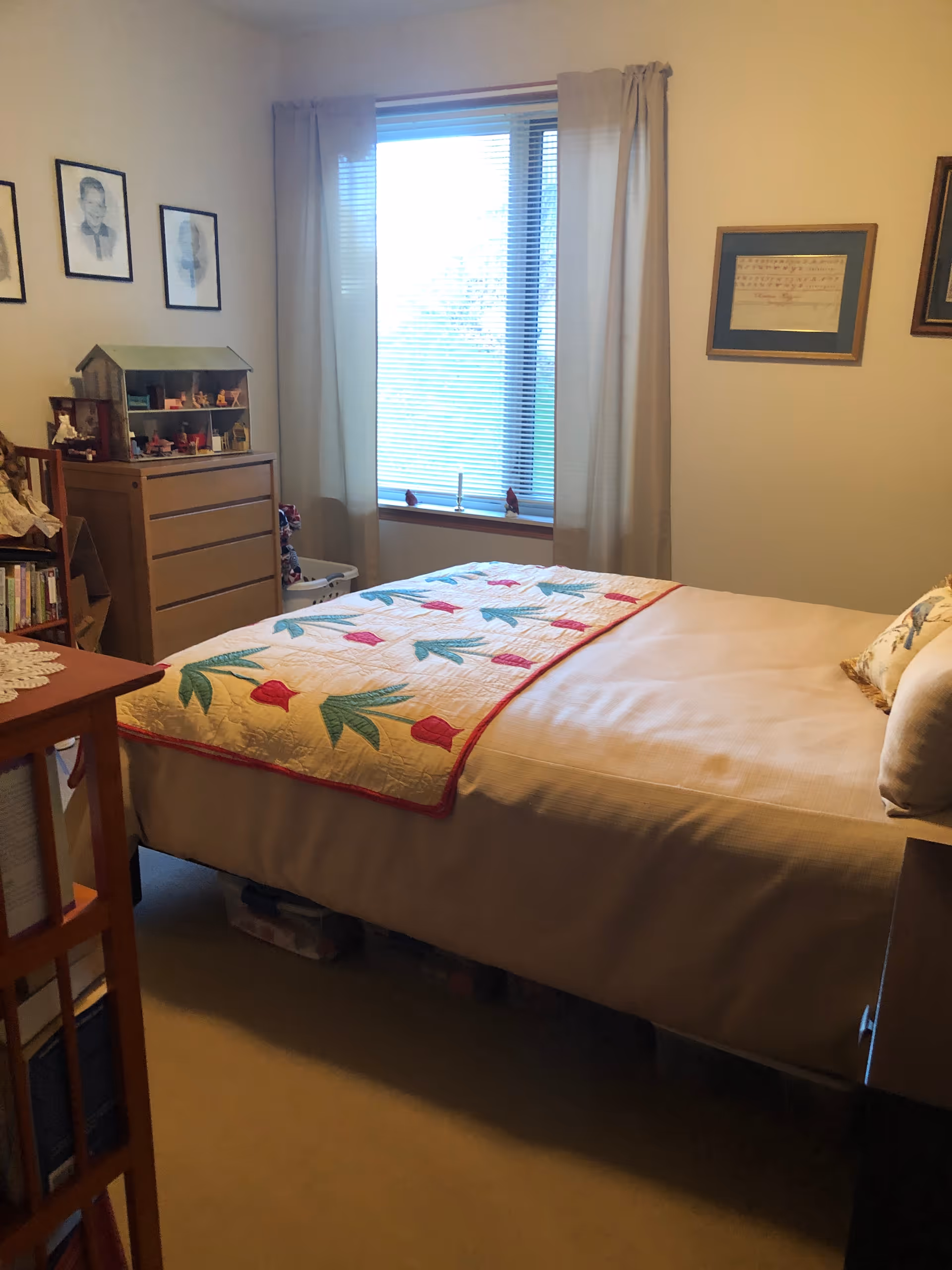 A cozy bedroom with a bed covered in a beige bedspread and a quilt with red and green leaf patterns. There is a window with closed blinds and beige curtains letting in natural light. A wooden dresser with a dollhouse on top and framed pictures on the walls are visible. A bookshelf with books and a doll is also present.