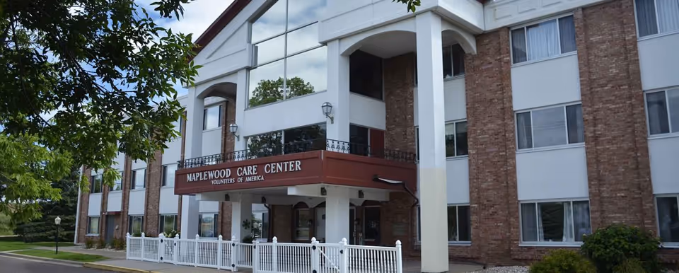 Front exterior of the Maplewood Care Center showing the entrance canopy with columns, brick facade, windows, and a white picket fence.