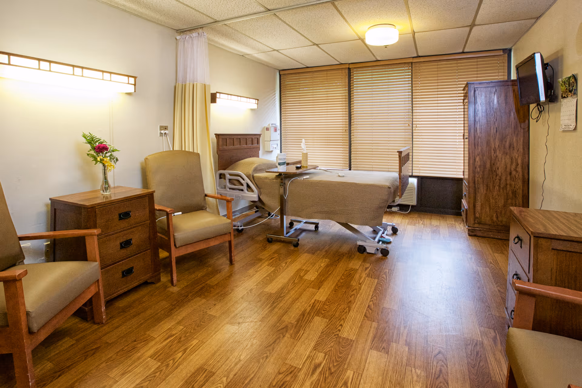 A well-lit patient room with a hospital bed covered in a beige blanket, two beige armchairs, wooden furniture including a nightstand with a vase of flowers, a dresser, and a wall-mounted TV. The room has wooden flooring and large windows with closed blinds.