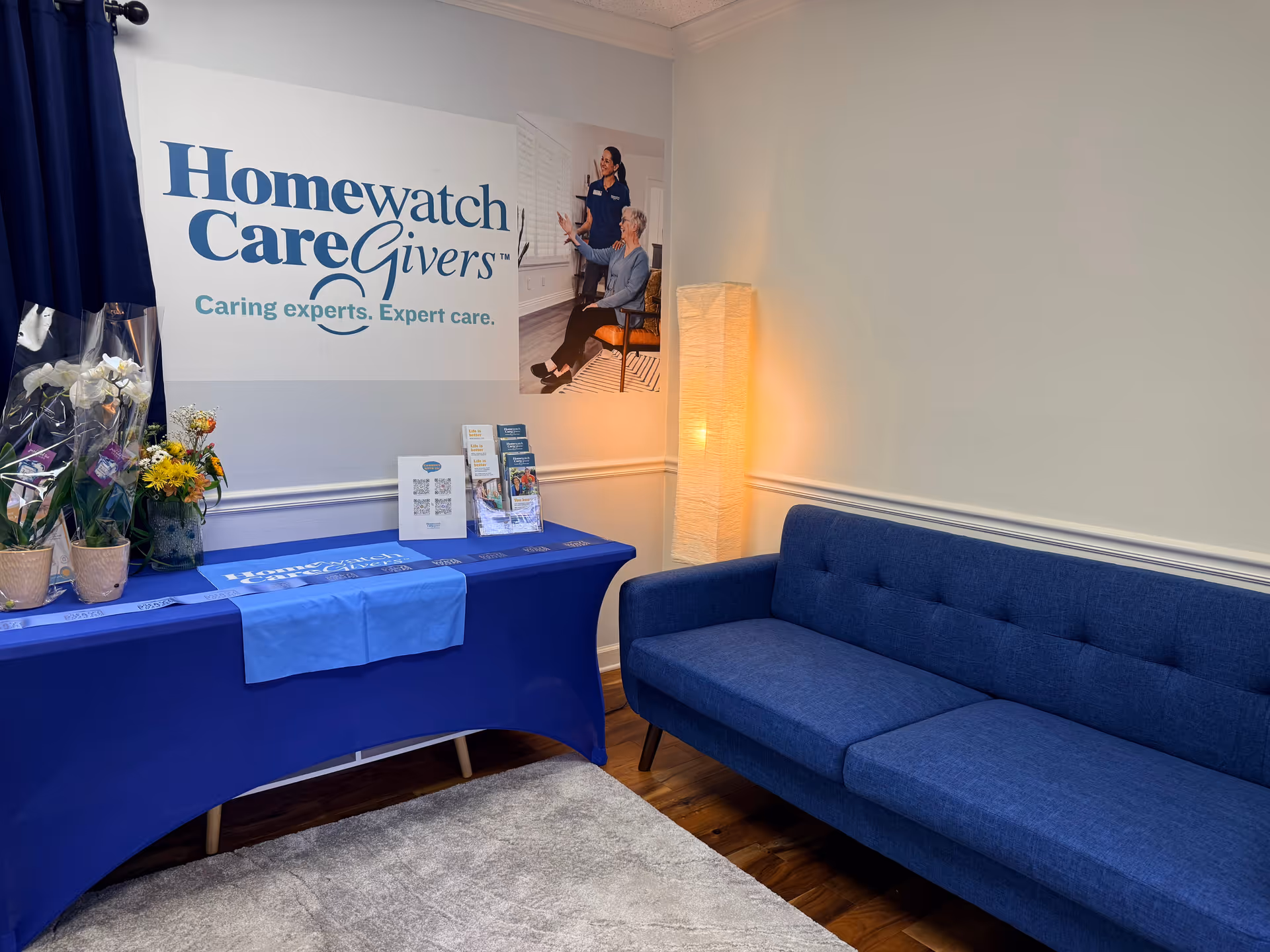 Interior lobby area with Homewatch CareGivers signage, blue sofa, plants, and brochures.