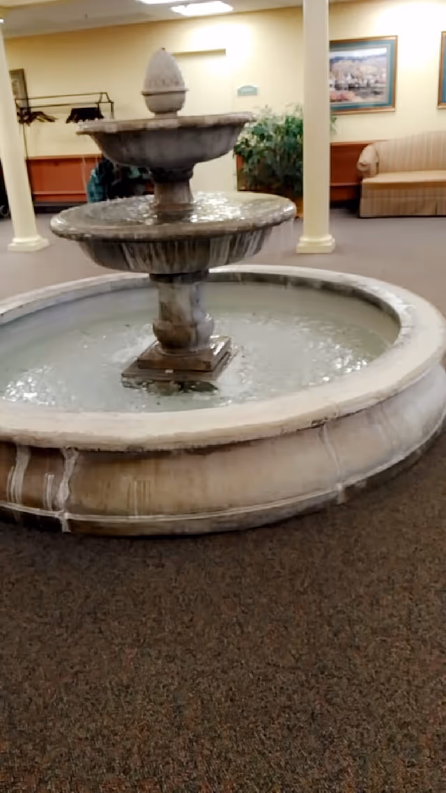 Indoor lobby fountain with seating, paintings, and potted plants.