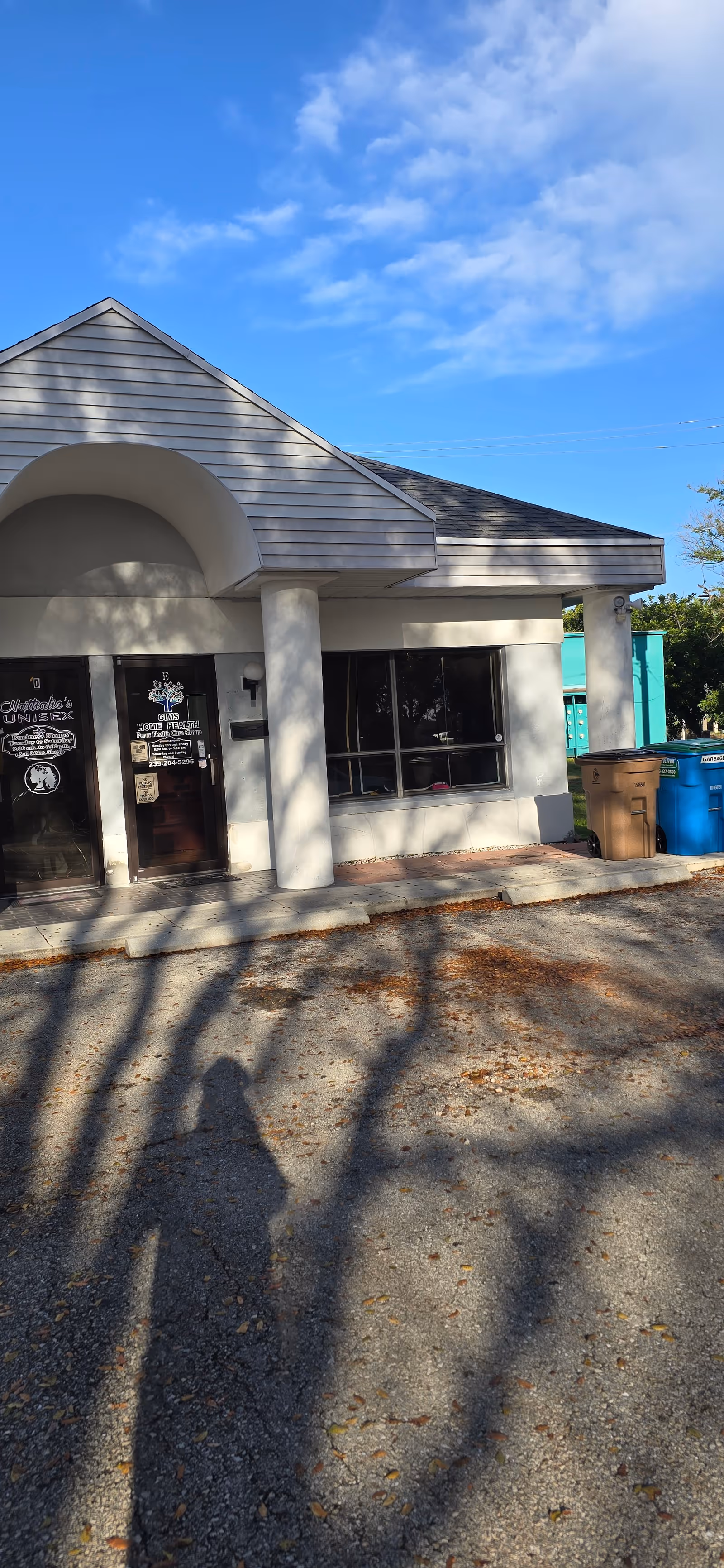Small pale building with arched entrance, glass windows, and trash bins outside under blue sky.