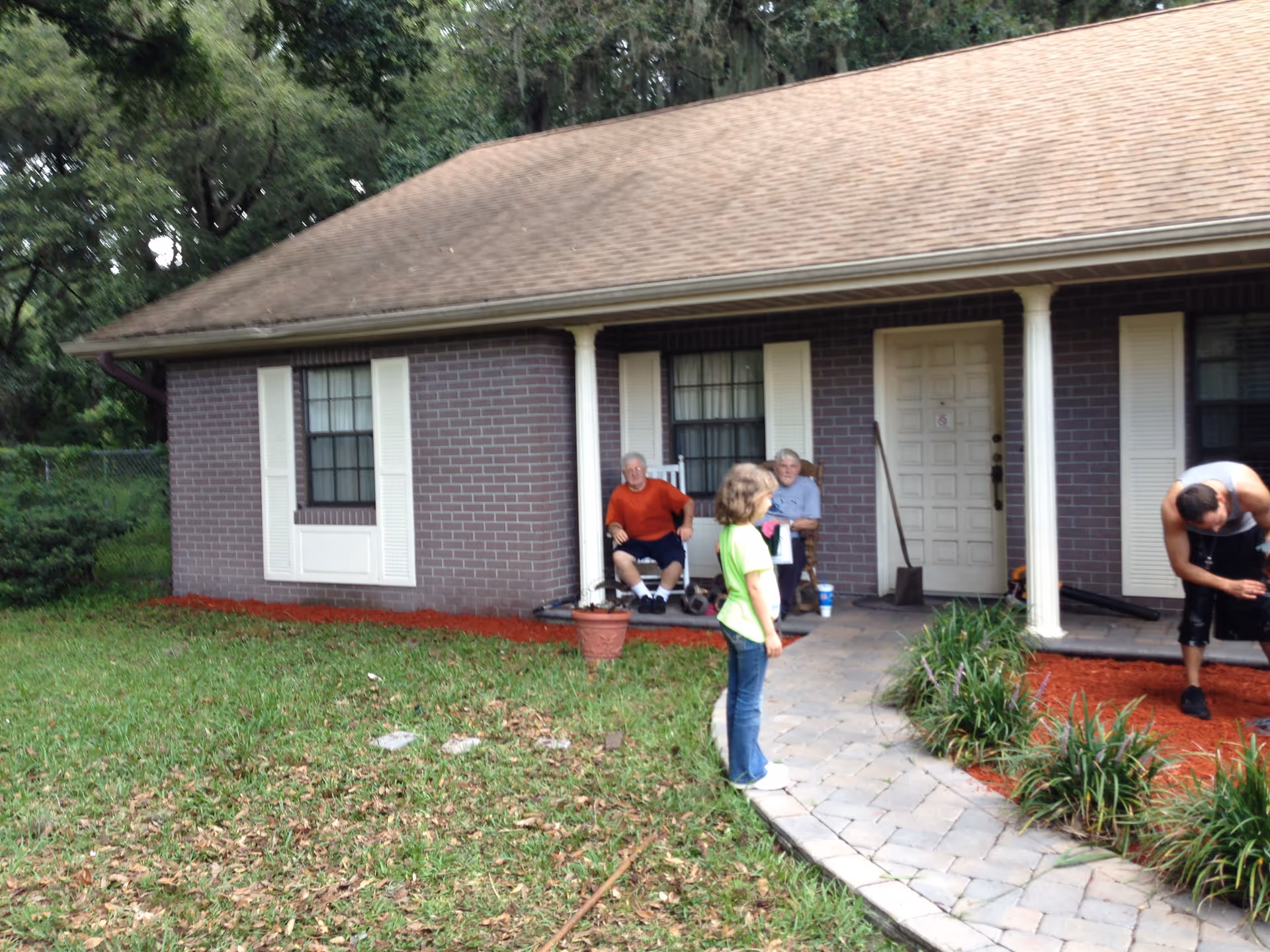 Front of a single-story brick house with a covered porch where several people stand and sit near a paved walkway and lawn.