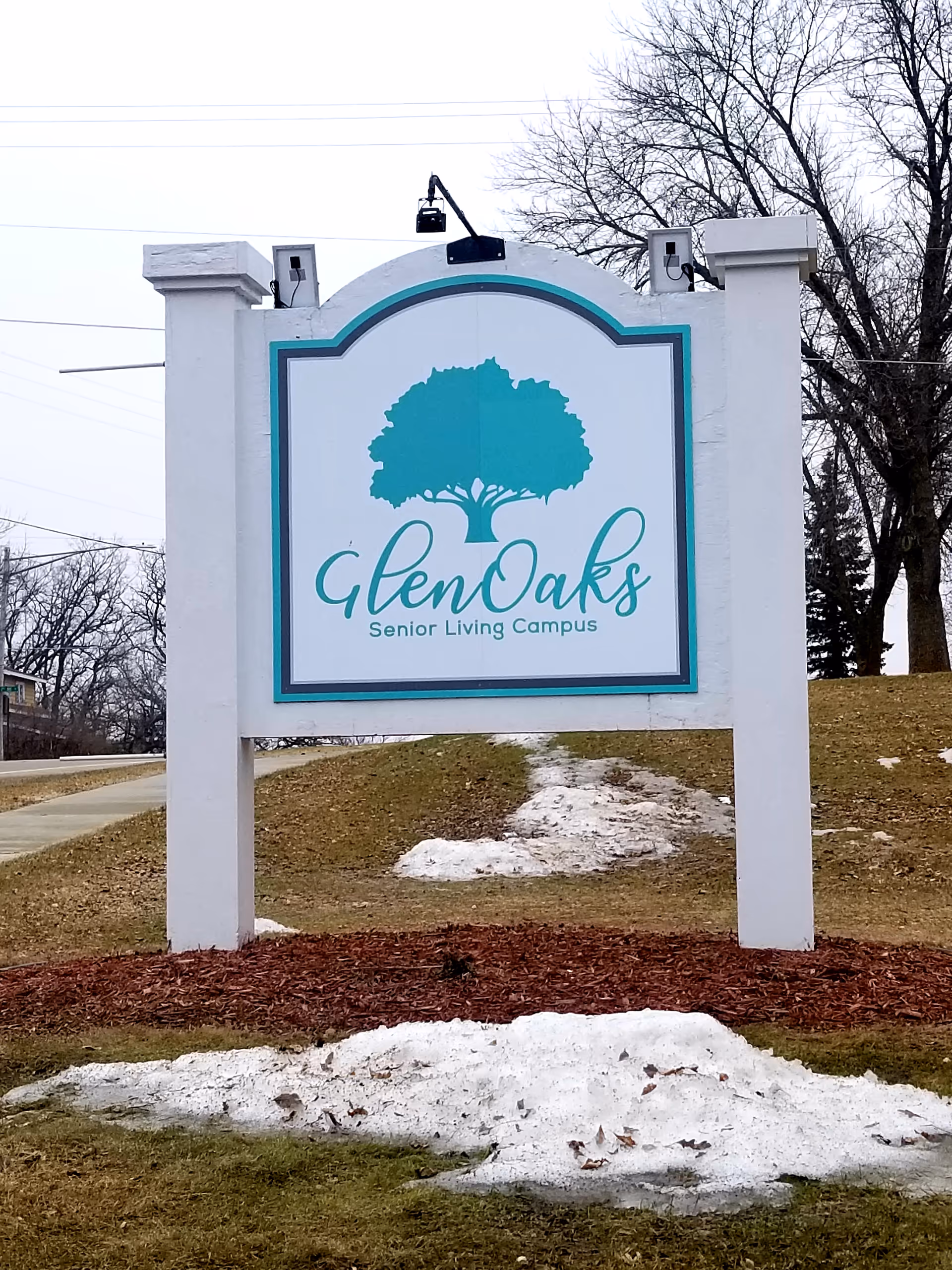A large white sign reads GlenOaks Senior Living Campus outdoors with teal tree logo.