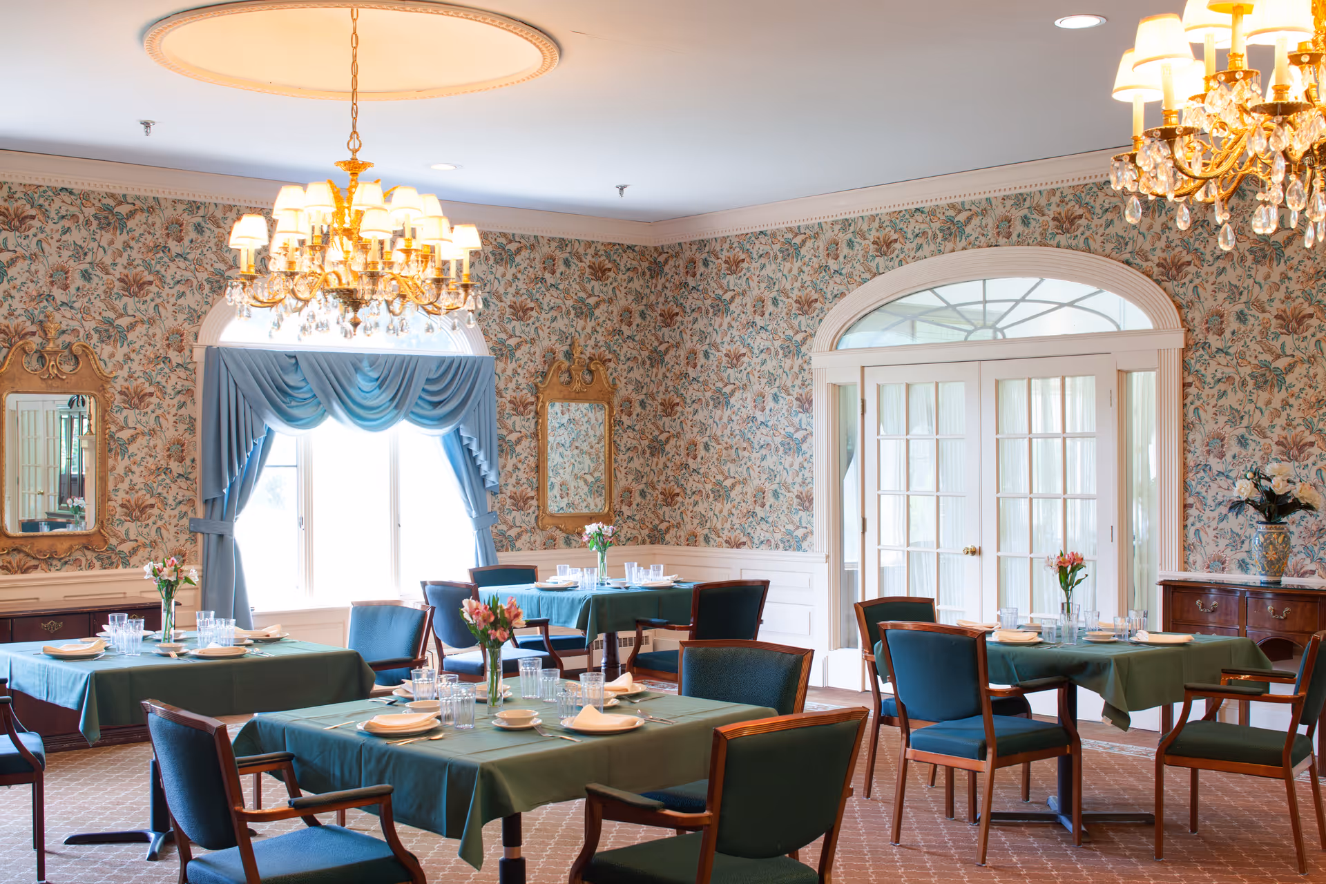 A dining room with several tables covered in green tablecloths, each set with plates, glasses, and napkins. The room features floral wallpaper, two ornate mirrors, blue drapes over a window, and a chandelier hanging from the ceiling. There are wooden chairs with blue cushions around the tables.