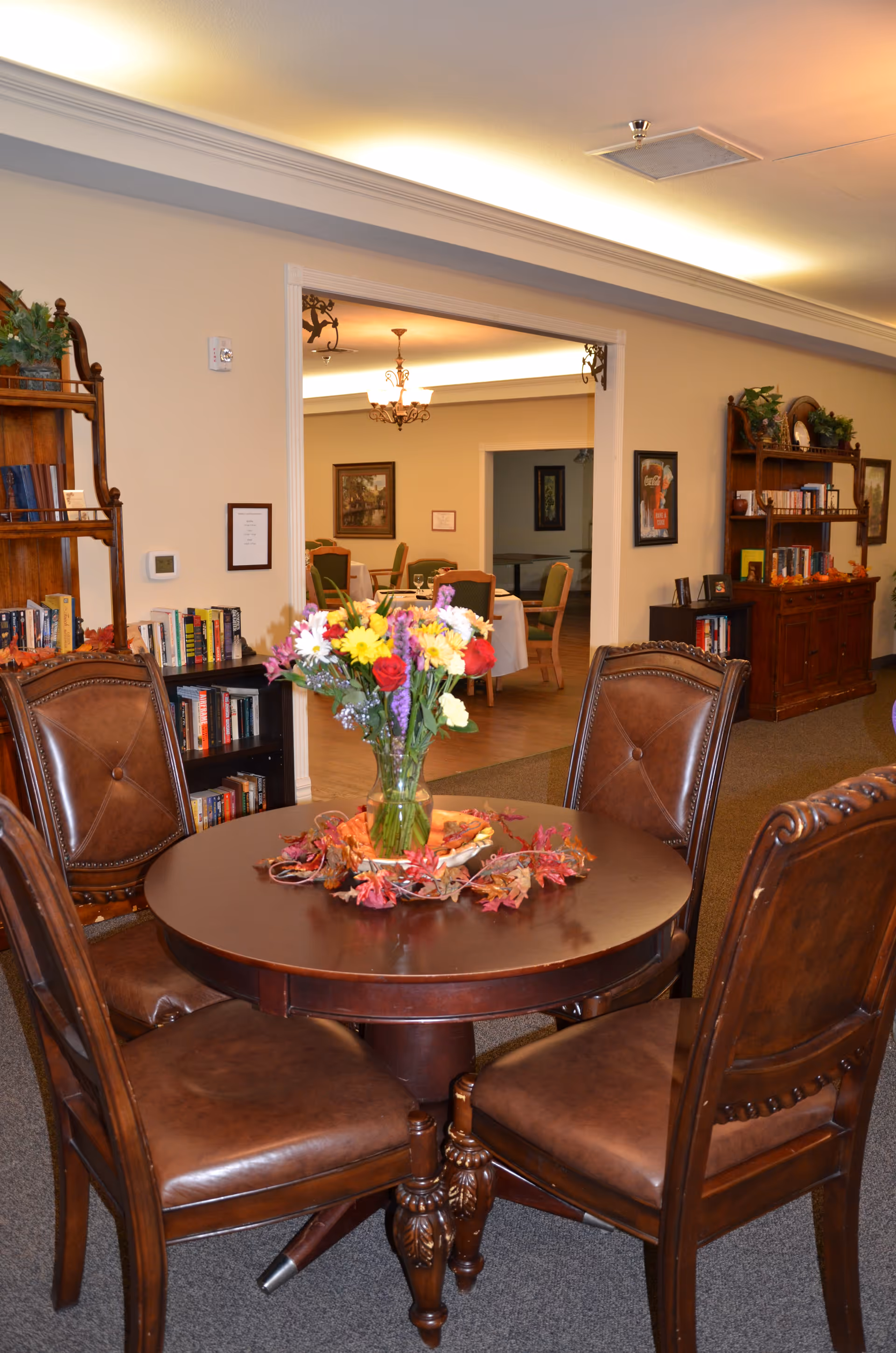 A cozy interior room with a round wooden table surrounded by four brown leather chairs. A vase with a colorful bouquet of flowers sits in the center of the table, decorated with autumn leaves. In the background, there are bookshelves filled with books and a view into another room with more chairs and tables, illuminated by chandeliers.