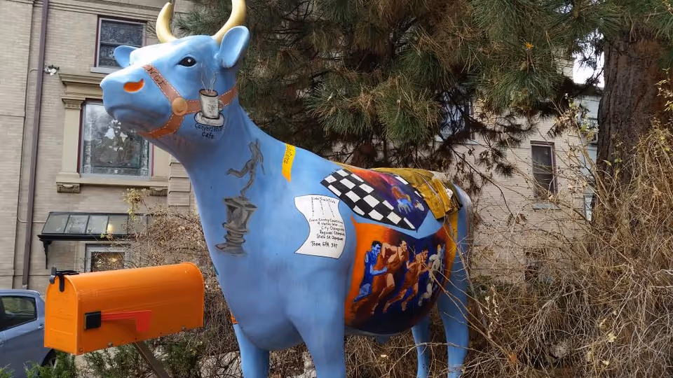 A large blue decorated cow sculpture stands outside a brick building near trees.