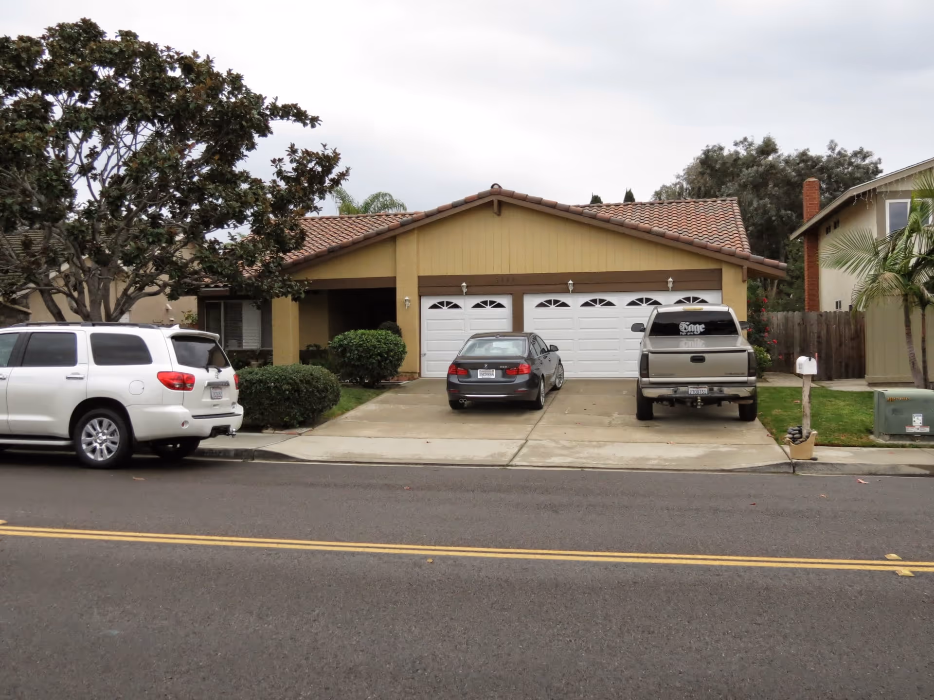 Front exterior view of a single-story house with a tiled roof, three white garage doors, and a driveway with three parked vehicles. There are bushes and trees around the house, and a mailbox on the right side near the street.