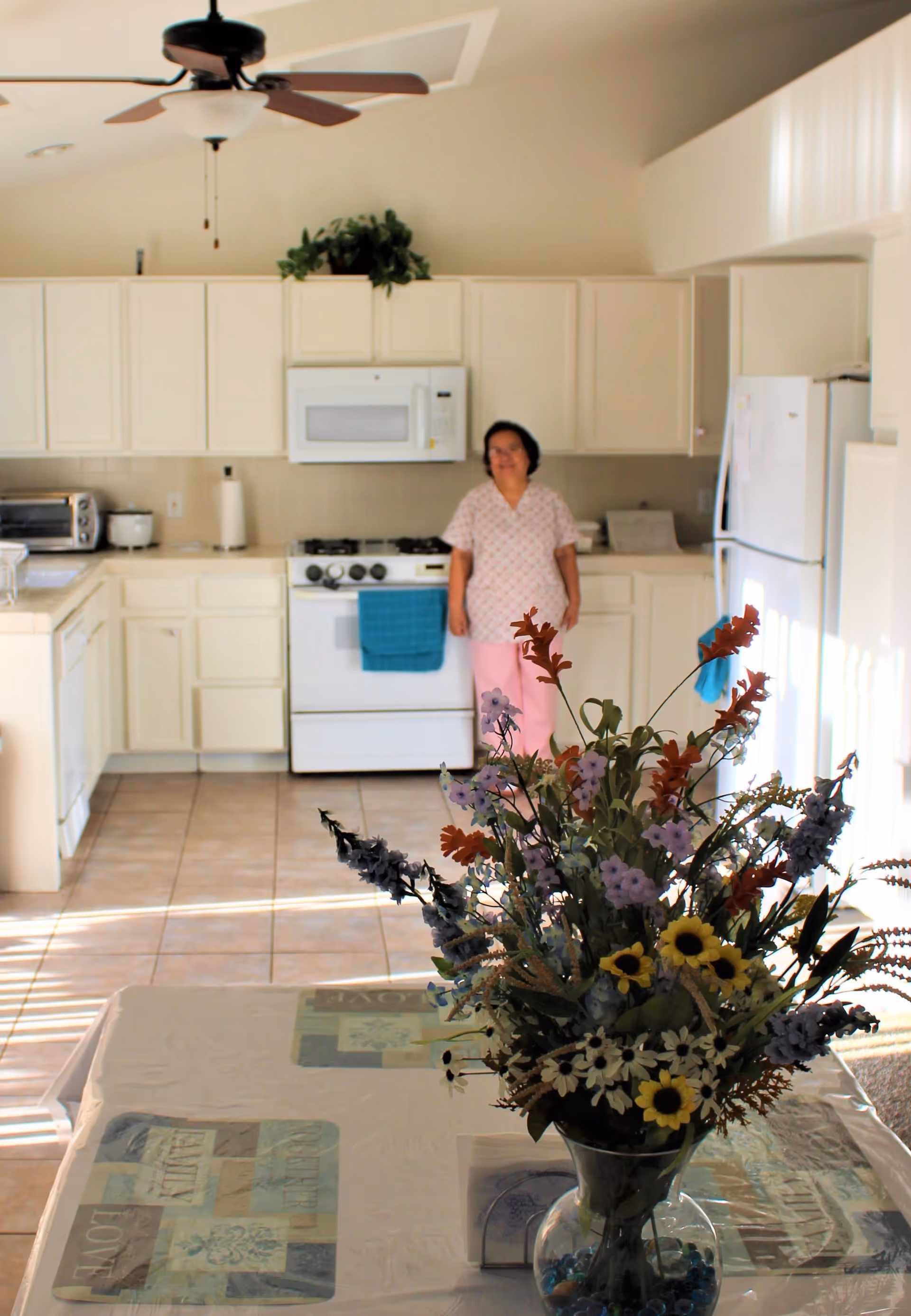 Sunlit kitchen with white cabinets and appliances, a vase of flowers on a table in the foreground, and a woman standing by the stove.