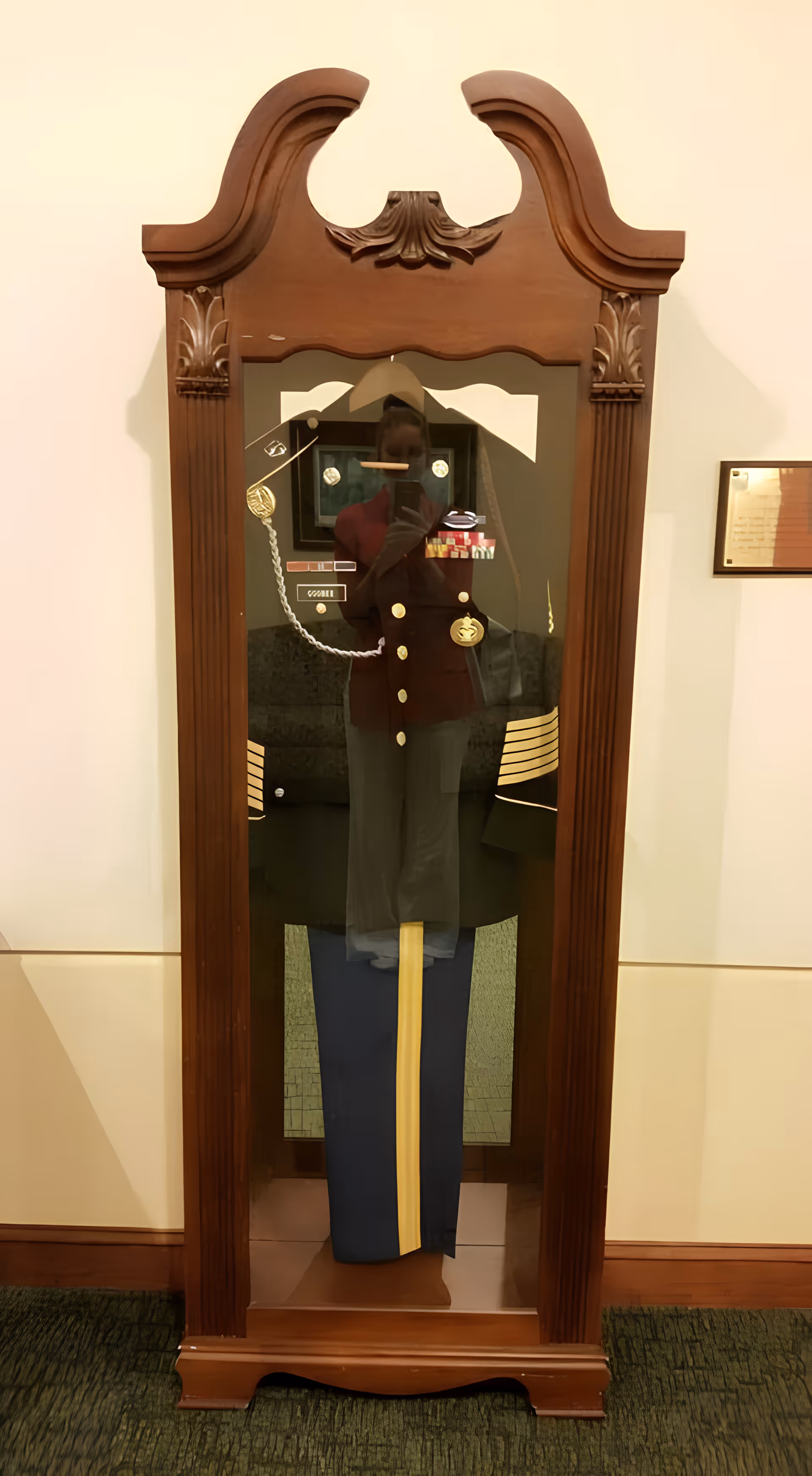 A wooden display case with a glass front containing a military dress uniform with medals and insignia. The case has ornate carved details at the top and is placed against a light-colored wall with a framed certificate or plaque visible on the right side.