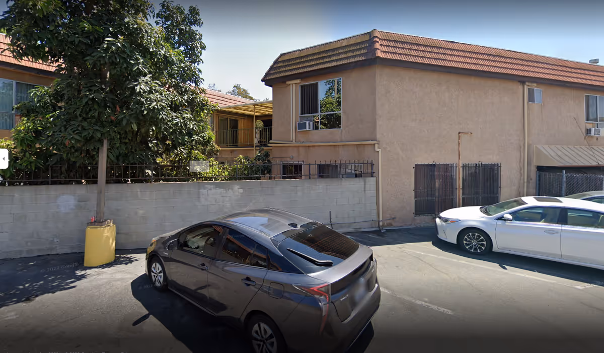 Parking lot with two cars in front of a two-story stucco building, a cinderblock wall, metal security gates, and a large tree.