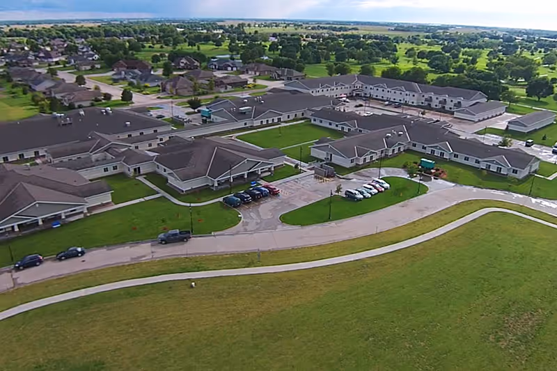 Aerial view of Good Samaritan Society - Grand Island Village facility showing multiple connected buildings with gray roofs, surrounded by green lawns and trees. Several cars are parked in front of the buildings along curved driveways and parking areas.