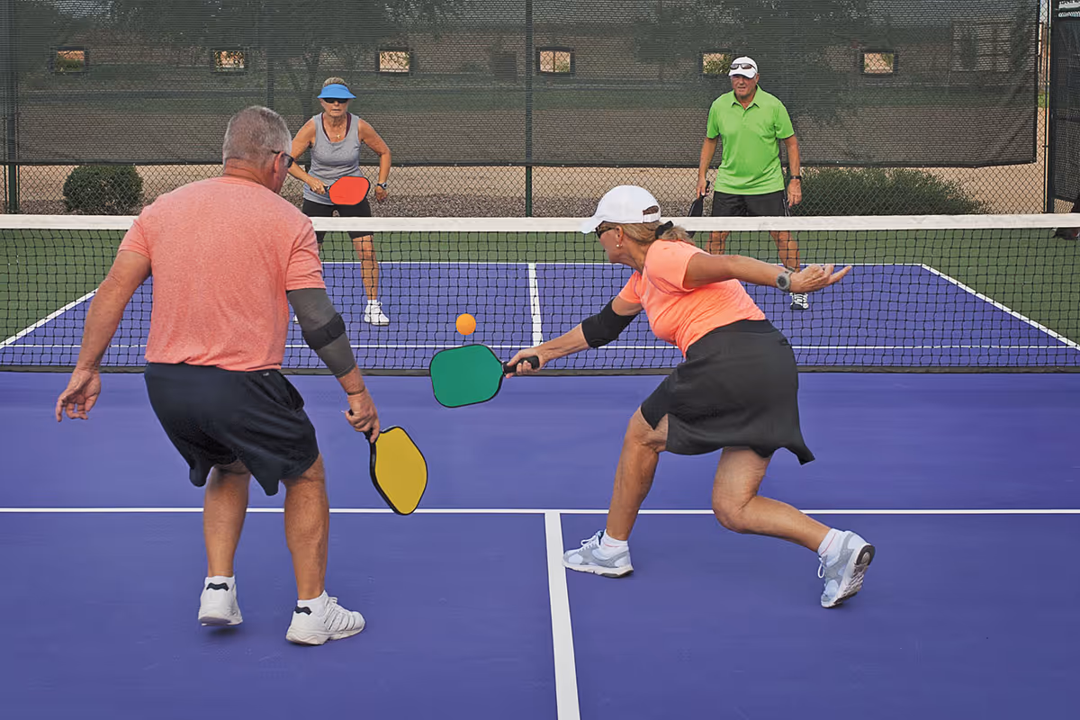 Four seniors playing pickleball on a purple court outdoors, two on each side of the net, actively engaged in the game with paddles and an orange ball.