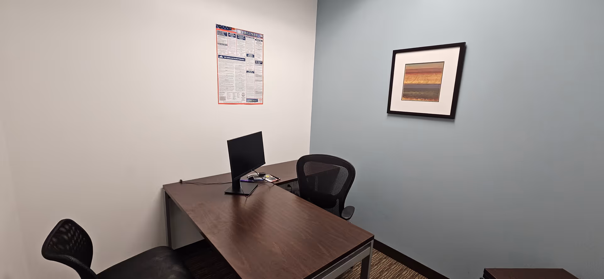A small office with a desk, computer monitor, chair, and framed artwork on walls.
