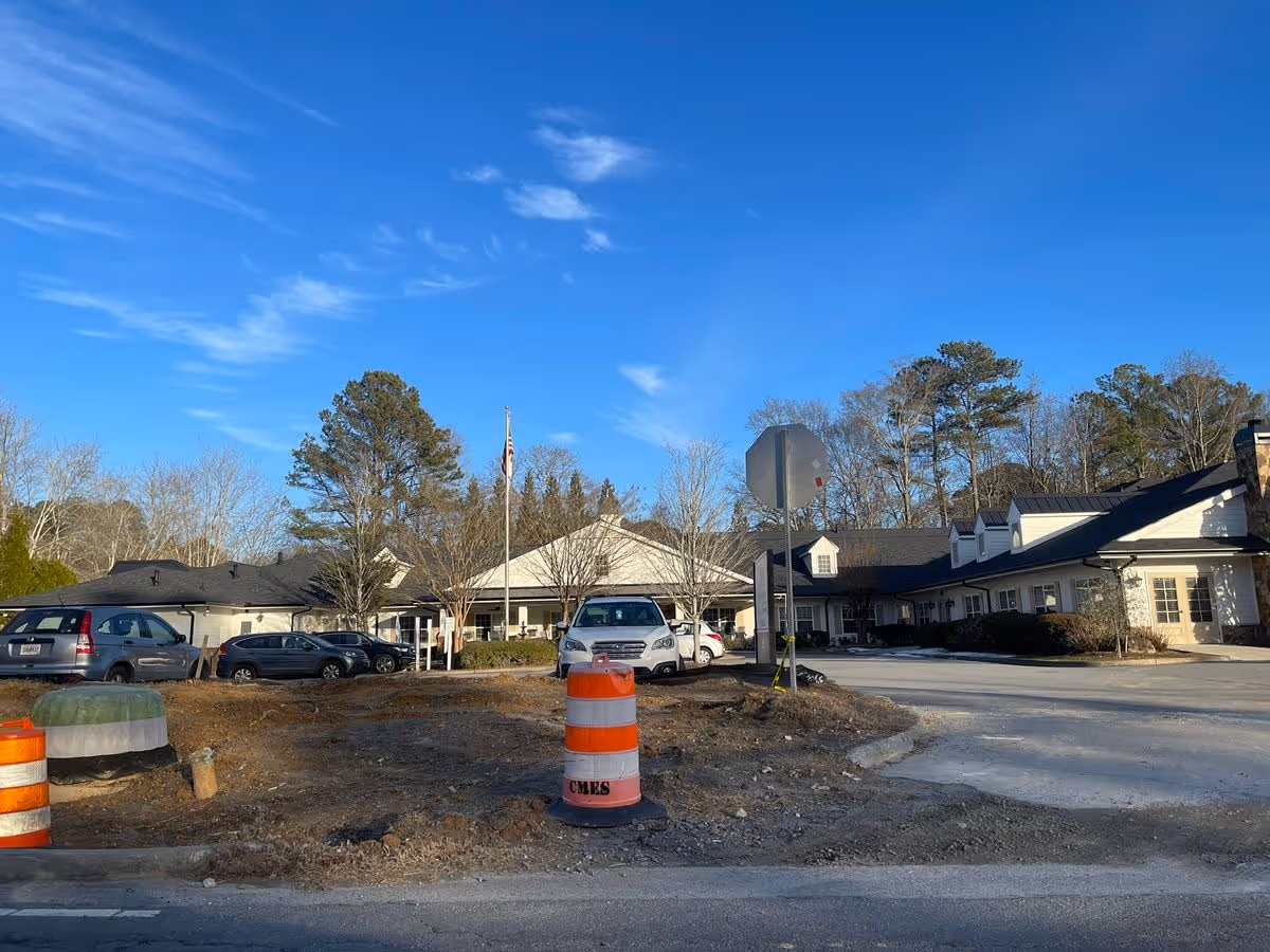 Exterior front view of a single-story senior living building with parked cars, traffic barrels, and a clear blue sky.