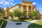 Sunlit tan three-story Tesoro Senior Apartments building with trees, manicured lawn, and curved driveway.