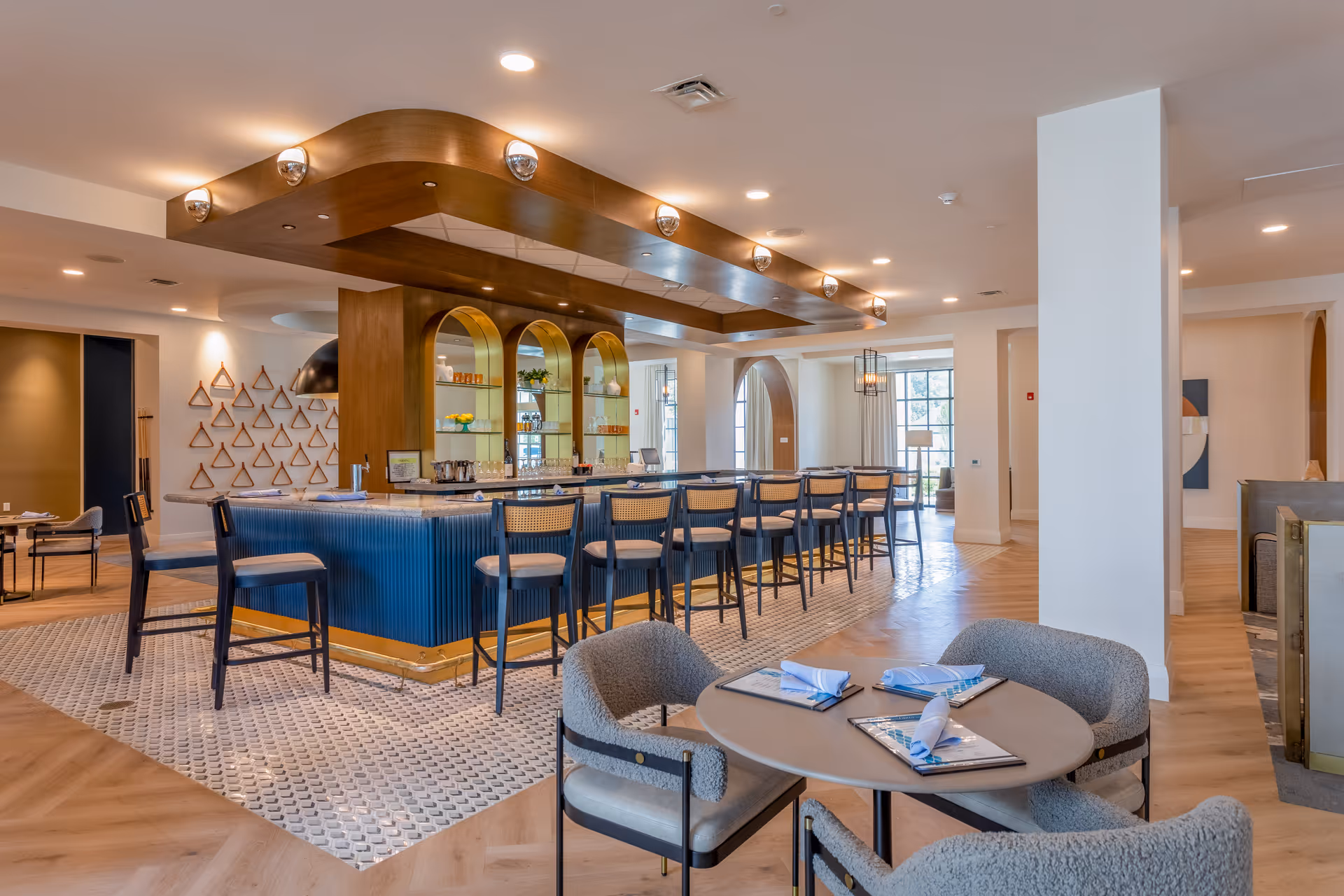 Modern communal bar and lounge area with a long blue-tiled counter, high stools, and round tables with upholstered chairs in a bright interior.