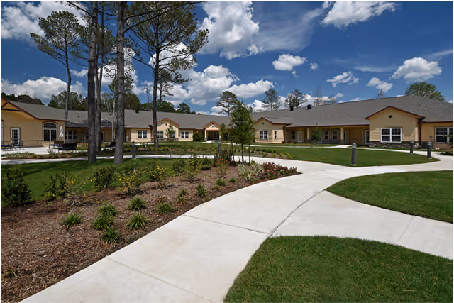 Wide view of the exterior of Ella Springs Senior Living Community building with a curved concrete walkway, landscaped garden beds, green grass, and tall pine trees under a partly cloudy blue sky.