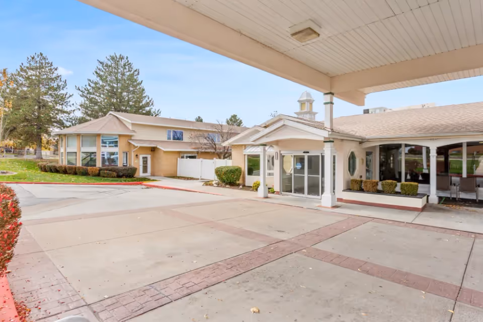 Exterior view of The Beaumont Assisted Living and Memory Care facility showing the entrance with a covered drop-off area, landscaped bushes, and trees in the background under a clear sky.