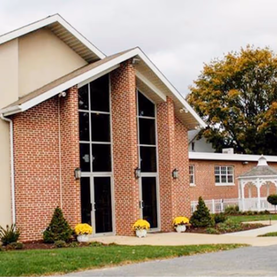 Brick facility front with tall glass doors, yellow potted flowers, and landscaped surroundings.