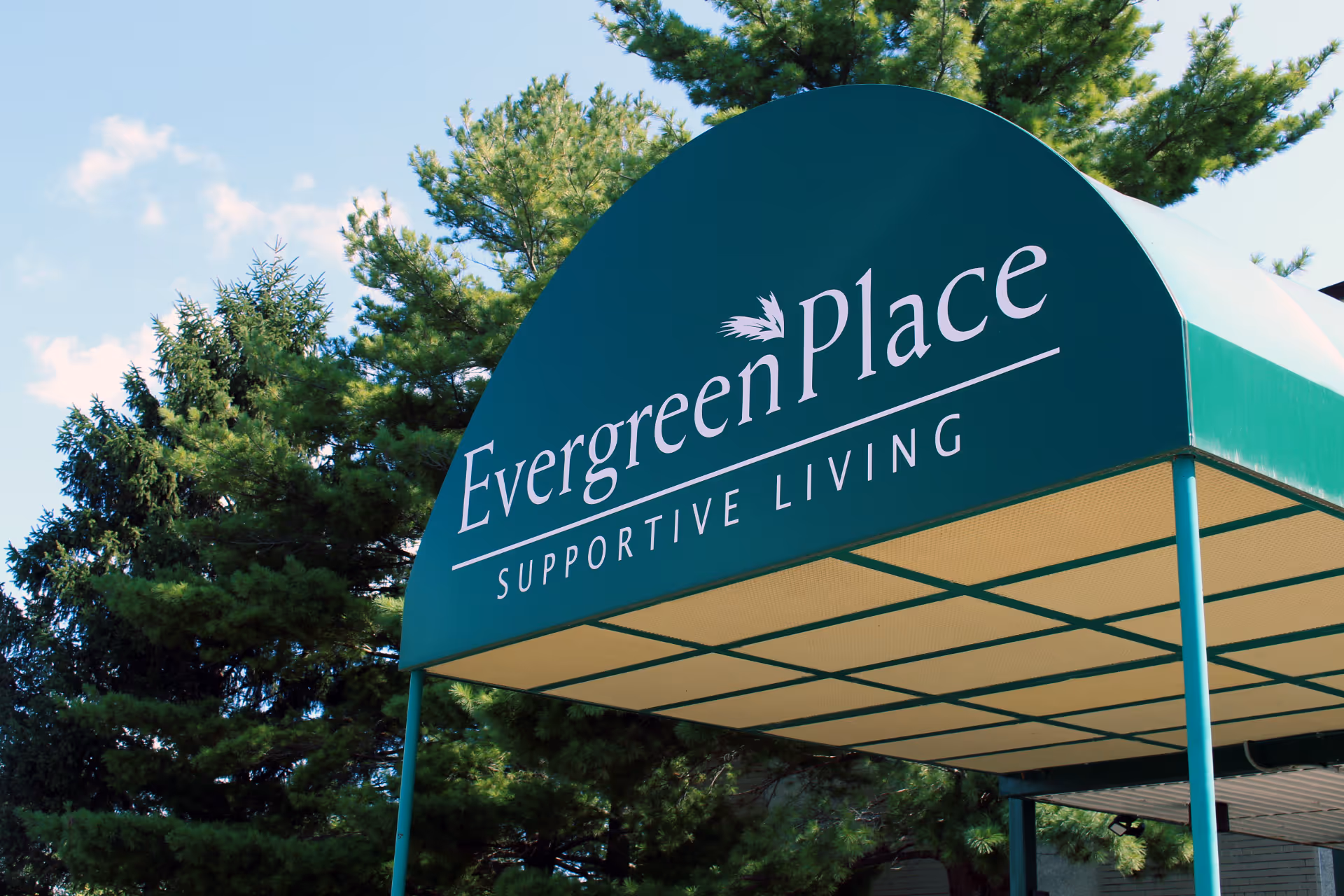 Green canopy with white text reading 'Evergreen Place Supportive Living' in front of tall green pine trees under a blue sky with some clouds.