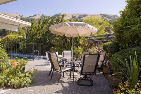 Outdoor patio with table, chairs, umbrella, and greenery surrounding a fenced pool area.