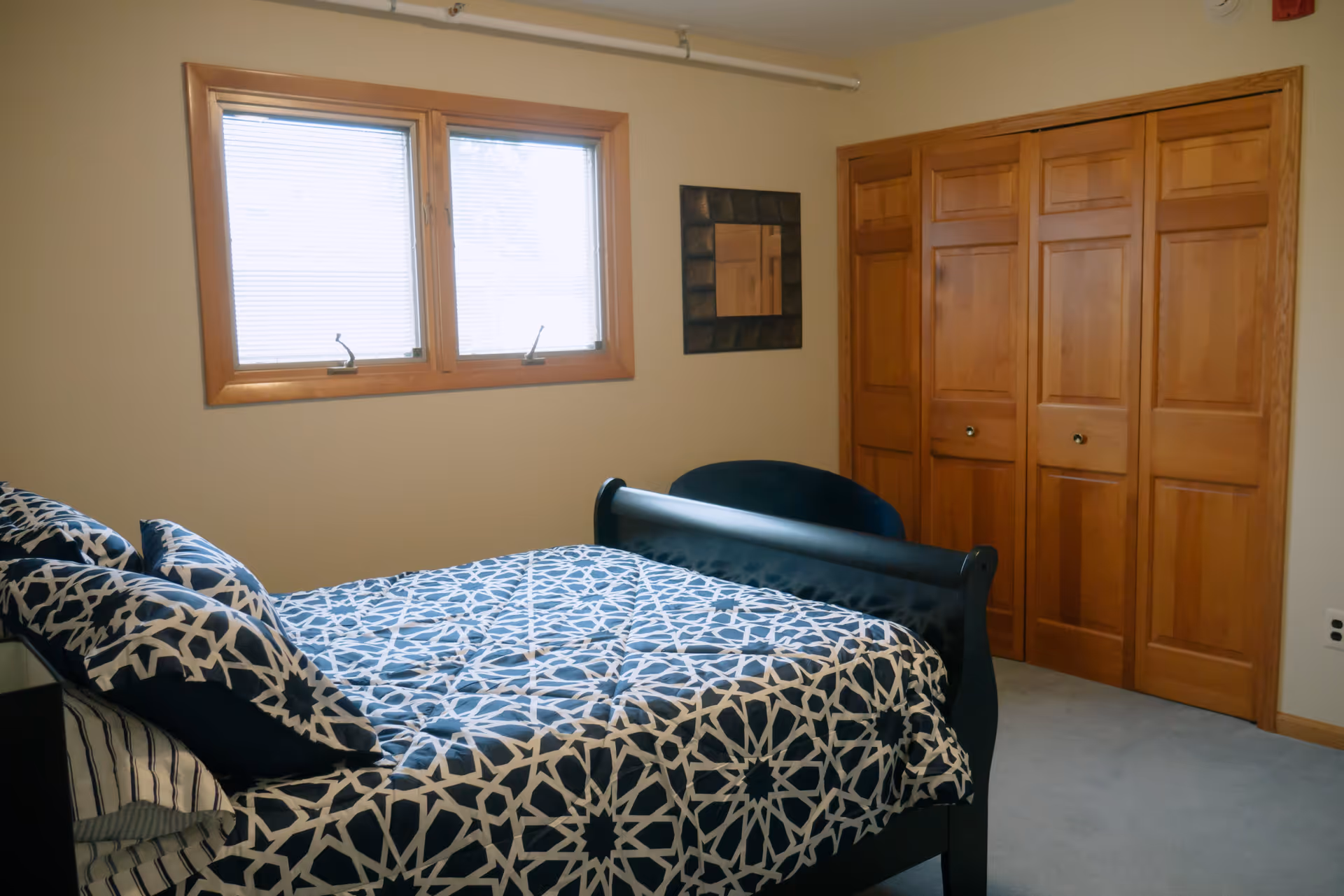 A bedroom with a bed covered in a blue and white patterned comforter and matching pillows. There is a window with blinds on the wall behind the bed, a dark chair next to the bed, a square decorative mirror on the wall, and a wooden closet with four paneled doors.