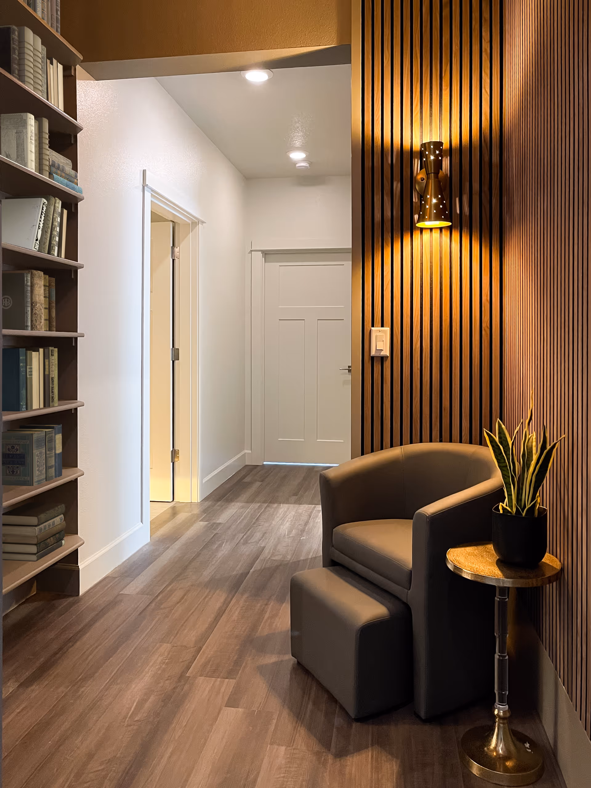 Cozy interior hallway seating area with a gray armchair and ottoman next to a small table and potted plant, wood slat accent wall, bookshelf, and a closed door down the hall.
