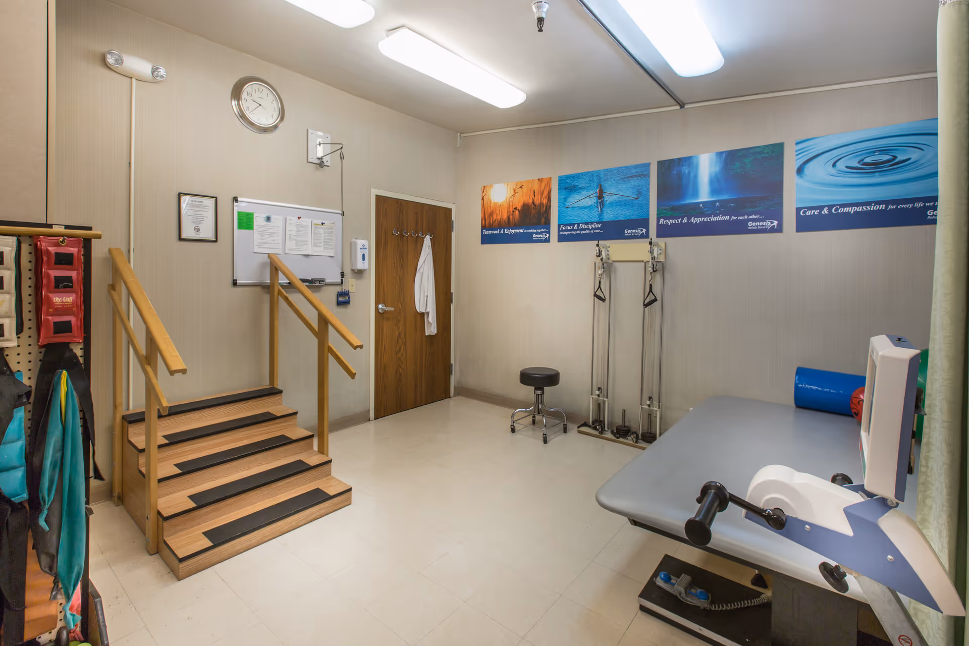 A rehabilitation or physical therapy room with wooden stairs for exercise, exercise equipment including pulleys and a pedal machine, a stool, and motivational posters on the wall. There is a wooden door with a white coat hanging on a hook, a clock, and a bulletin board with papers on the wall.