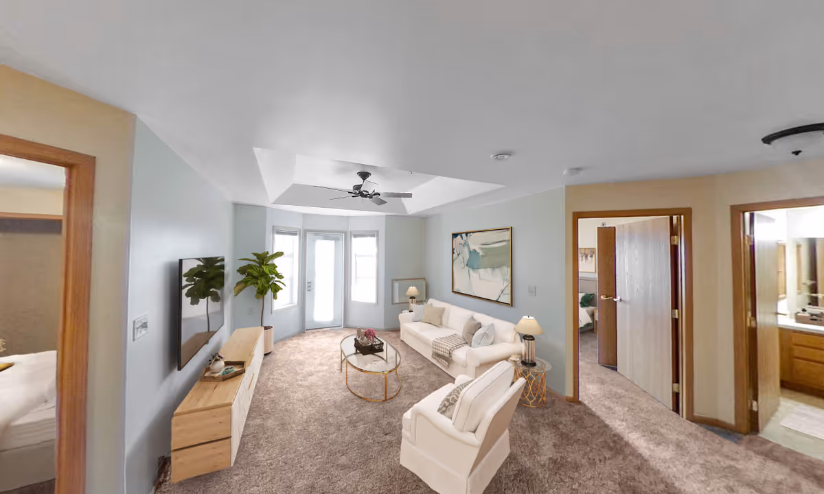 Bright furnished living room with a sofa, armchair, coffee table, TV console, ceiling fan, and doorways to adjacent rooms.