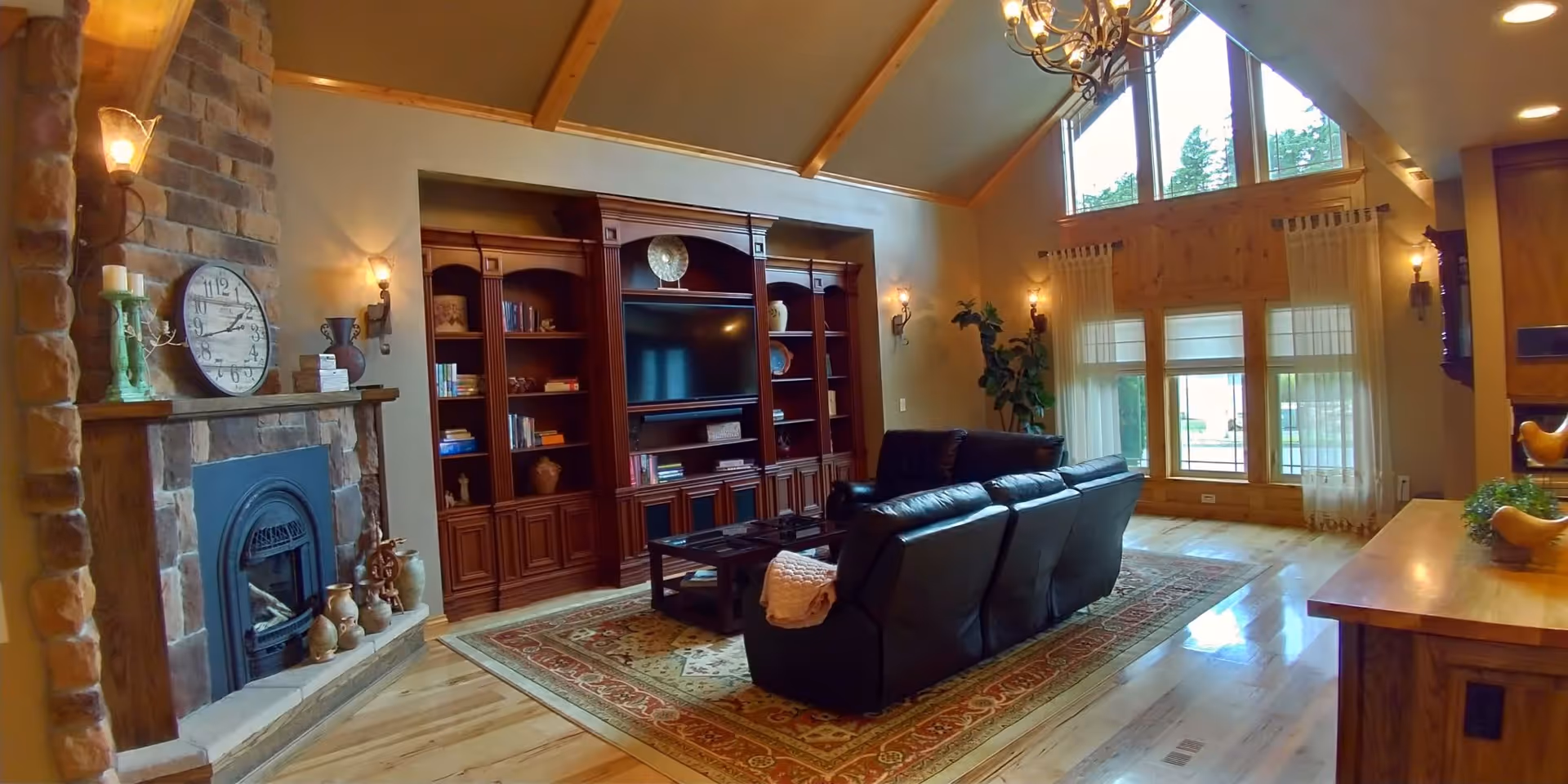 Spacious living room with vaulted ceiling, stone fireplace, built-in wooden shelving, leather sofa and large windows.