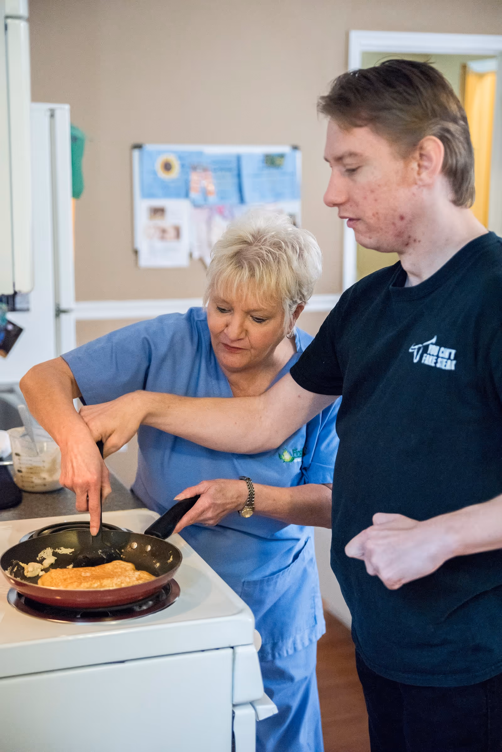 An elderly resident and a staff member cook together in a home-like kitchen.