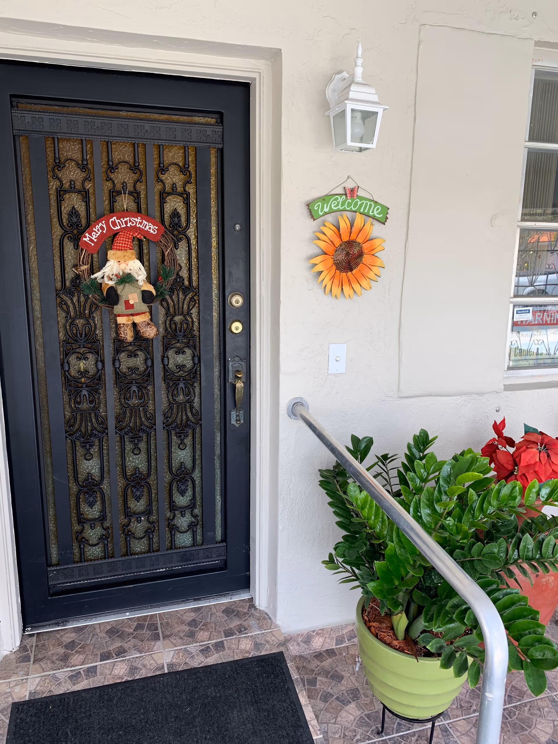 Front entrance with ornate iron door, Christmas wreath, sunflower welcome sign, and potted greenery.